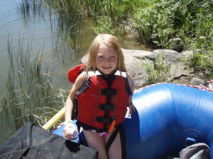 a girl in a red lifejacket sits on a blue boat smiling and waiting to get rafting on her hells canyon rafting trip